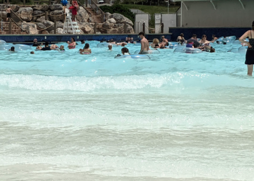 A crowded wave pool with people floating on blue inflatables, enjoying the water and waves on a sunny day.
