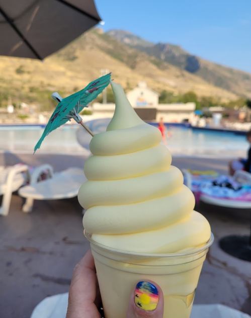 A hand holding a soft-serve ice cream cone with a small umbrella, set against a scenic outdoor backdrop.