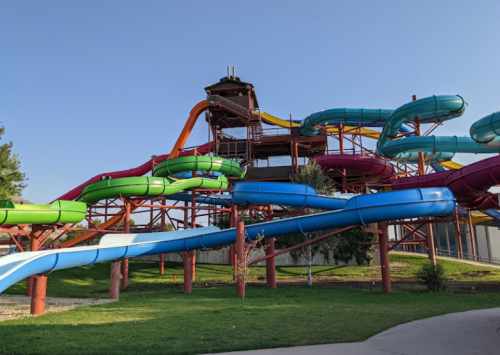 Colorful water slides twist and turn on a metal structure, surrounded by green grass and a clear blue sky.