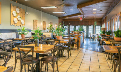 A cozy restaurant interior with wooden tables, plants, and warm lighting, featuring a rustic design and spacious layout.