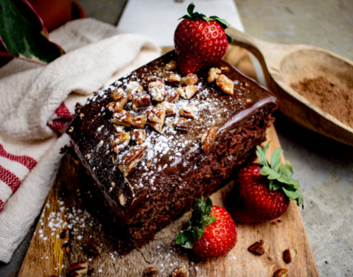 A chocolate cake topped with nuts and strawberries, dusted with powdered sugar, on a wooden board.