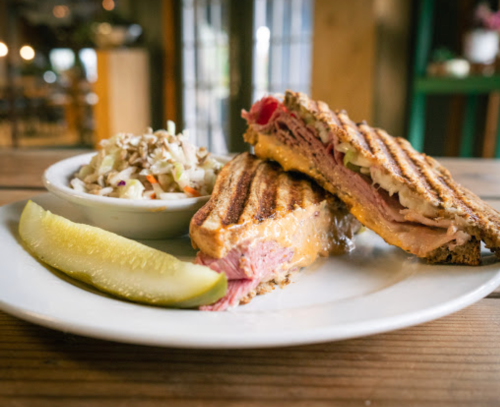 A grilled sandwich with ham and cheese, served with coleslaw and a pickle on a white plate.