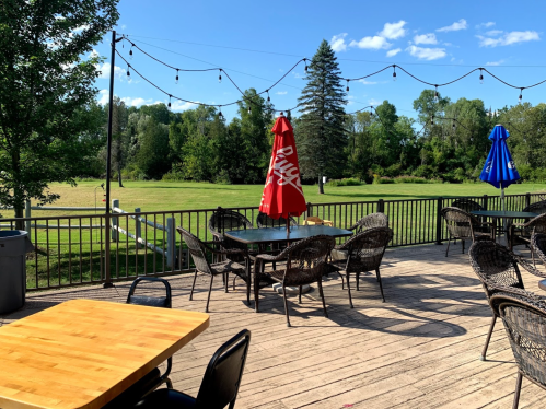 Outdoor patio with tables and chairs, featuring red and blue umbrellas, overlooking a green field and trees.