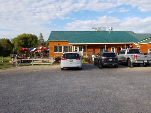 A rustic building with a green roof, surrounded by cars and outdoor seating under umbrellas on a gravel lot.