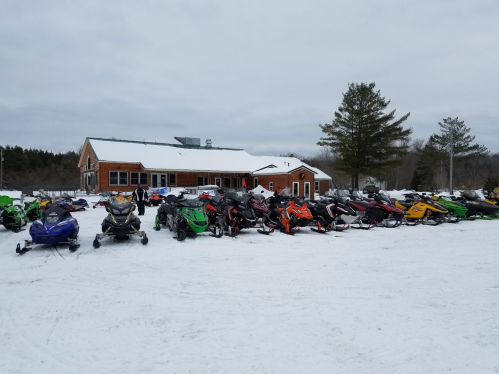 A line of colorful snowmobiles parked in the snow outside a rustic lodge on a cloudy winter day.