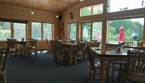Cozy wooden restaurant interior with tables, chairs, and large windows overlooking a green landscape.