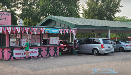 A colorful drive-in restaurant with a pink exterior, serving ice cream, surrounded by parked cars and trees.