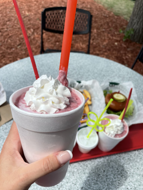 A hand holds a frosty drink topped with whipped cream and two straws, with food items on a table in the background.