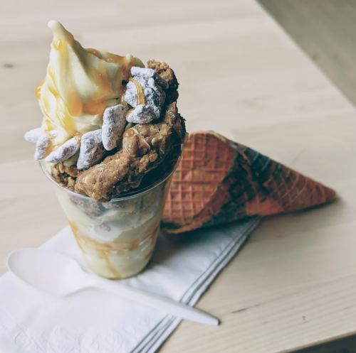 A dessert cup with soft serve, cookie pieces, and caramel drizzle, alongside a waffle cone on a napkin.