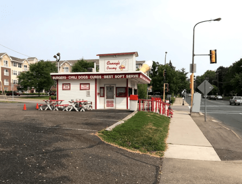 A small, red-and-white fast food stand with outdoor seating, offering burgers, chili dogs, and soft serve ice cream.