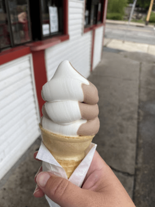 A hand holding a soft-serve ice cream cone with chocolate and vanilla swirls, in front of a building.