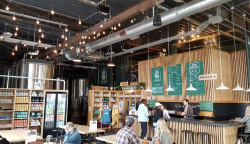A cozy brewery interior with wooden tables, shelves of merchandise, and patrons enjoying drinks under string lights.