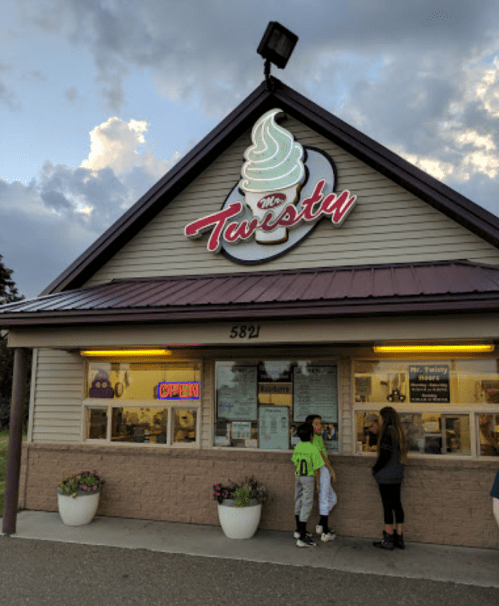 A small ice cream shop named "The Twisty" with a bright sign, featuring two customers at the window on a cloudy day.