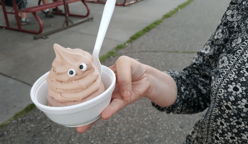 A person holds a cup of chocolate soft serve ice cream with googly eyes on top, set against a park background.