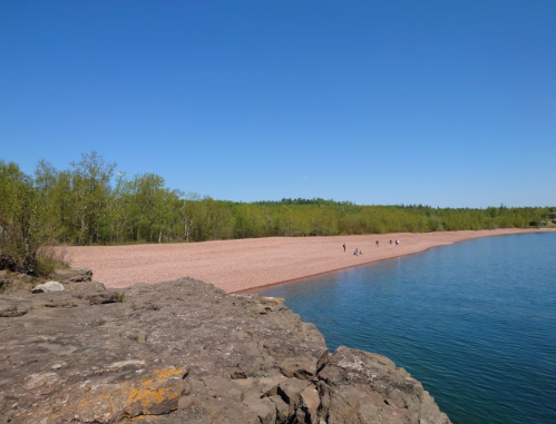 A sandy beach beside a calm lake, surrounded by green trees under a clear blue sky. People walk along the shore.