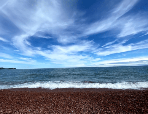 A serene beach scene with gentle waves lapping at a pebbly shore under a bright blue sky with wispy clouds.