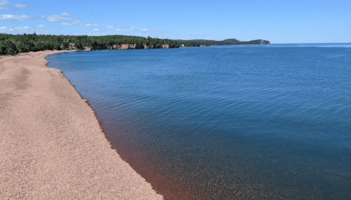 A serene beach with a sandy shore and calm blue water, surrounded by green trees and distant hills under a clear sky.