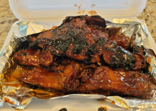 A takeout container with foil holding pieces of glazed, grilled meat, slightly charred and covered in sauce.