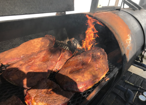 Smoked meats sizzling on a grill, with flames licking the side of the smoker.