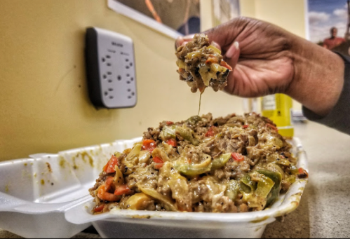 A hand reaches for a cheesy, meaty dish with peppers, served in a takeout container.