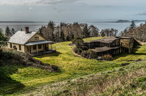 A scenic view of a yellow house and a large structure on a grassy hillside overlooking a calm body of water.