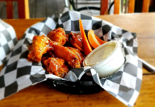 A basket of saucy chicken wings with carrot sticks and a small cup of dipping sauce, served on checkered paper.