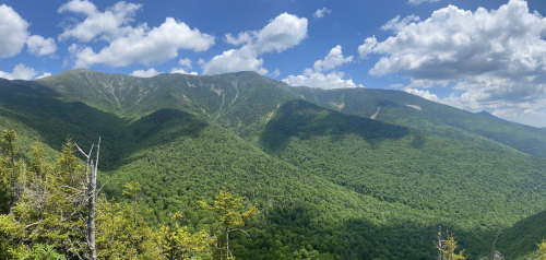 A panoramic view of lush green mountains under a blue sky with fluffy white clouds.