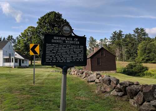 Historical marker for the birthplace of Daniel Webster, with a grassy area and buildings in the background.