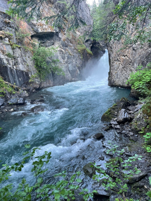 A rushing river flows through a narrow canyon, surrounded by rocky cliffs and lush greenery. Mist rises from the water.