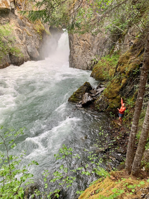 A person in an orange jacket stands by a rushing river, surrounded by lush greenery and rocky cliffs.