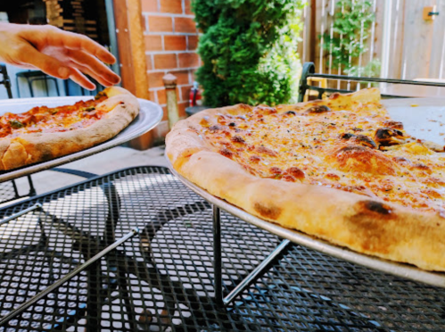A hand reaches for a slice of pizza on a table, with another pizza on a separate tray nearby.