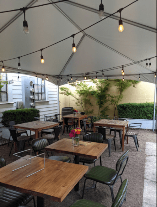 Outdoor dining area under a tent, featuring wooden tables, chairs, and string lights, with greenery in the background.