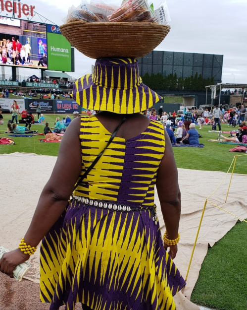 A person in a vibrant yellow and purple patterned dress carries a basket on their head at an outdoor event.