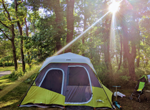 A green and gray tent set up in a wooded area, with sunlight streaming through the trees.