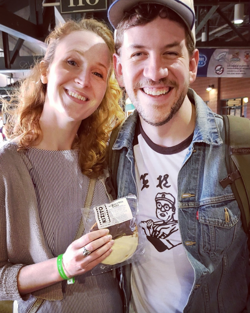 A smiling couple poses together at an event, holding a cookie. The woman has curly hair, and the man wears a denim jacket.