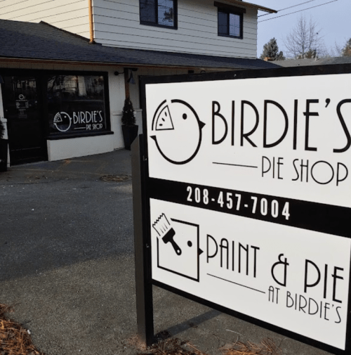 Sign for Birdie's Pie Shop featuring a logo and contact number, with a cozy building in the background.