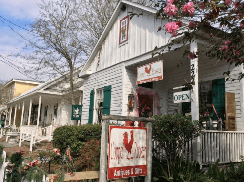 A quaint white building with a rooster sign, labeled "Copper Rooster Antiques & Gifts," surrounded by greenery and flowers.