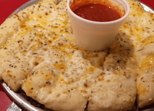 A cheesy pizza with golden crust and a cup of marinara sauce in the center, served on a round metal tray.