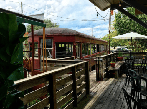 A vintage red train car is visible from a wooden deck with outdoor seating and greenery in the background.