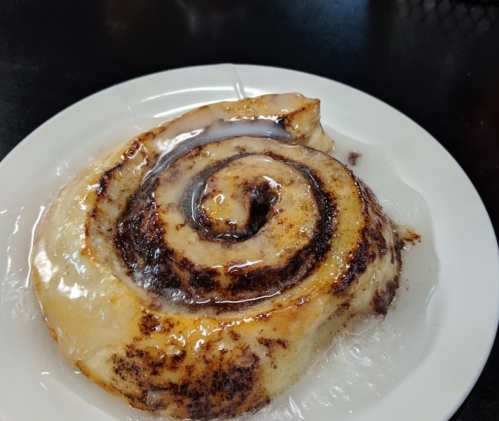 A freshly baked cinnamon roll on a white plate, drizzled with sweet icing.