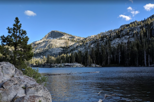 A serene lake surrounded by rocky shores and tall trees, with a mountain peak in the background under a clear blue sky.