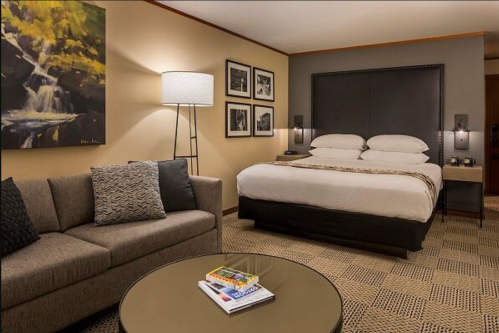 A cozy hotel room featuring a bed, sofa, coffee table, and artwork on the walls. Warm lighting creates a welcoming atmosphere.