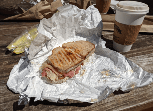 A grilled sandwich wrapped in paper, alongside a coffee cup and a couple of bananas on a wooden table.