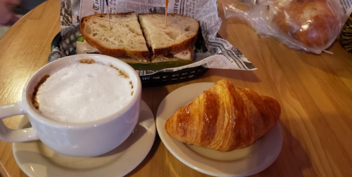 A cup of cappuccino, a sandwich on a plate, and a buttery croissant on a small dish, all on a wooden table.