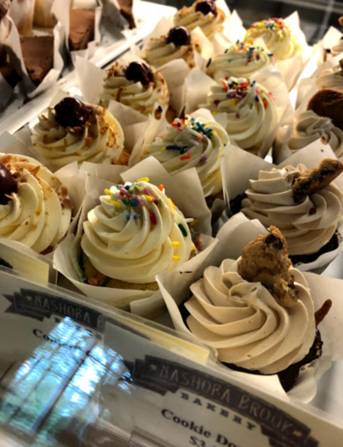 A display of colorful cupcakes with frosting, sprinkles, and cookie toppings in a bakery case.