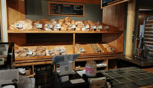 A bakery display with various bread and pastries on shelves, a cash register, and a drink tap in the foreground.