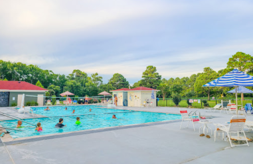 A sunny outdoor pool scene with people swimming, lounge chairs, and umbrellas under a blue sky.