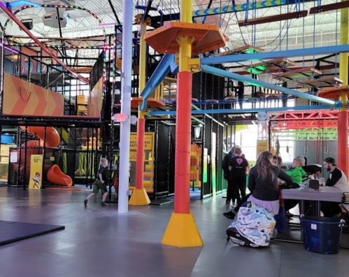 A colorful indoor play area with climbing structures, slides, and people socializing at tables.