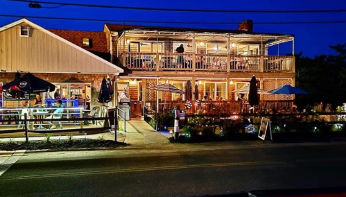 A cozy restaurant with outdoor seating, warmly lit at night, featuring a wooden exterior and umbrellas on the patio.