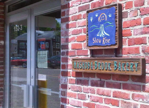 Sign for "Slow Rise" and "Hasswood Brook Bakery" on a brick wall next to a glass door.
